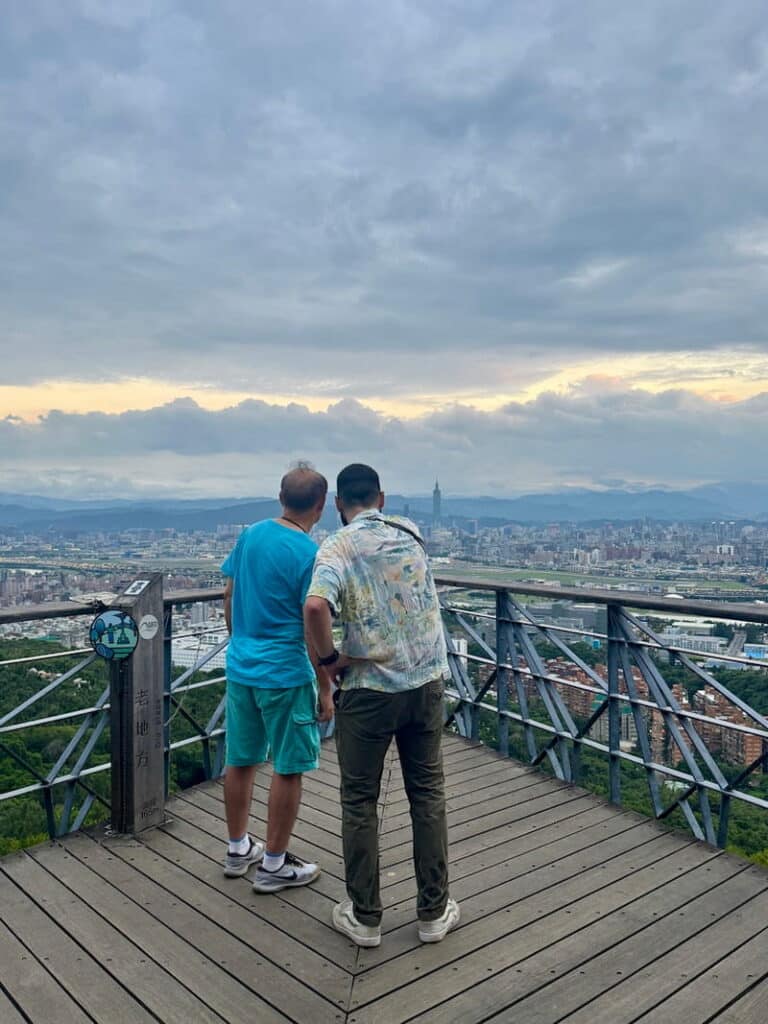 Two people standing on a wooden viewing platform at Lao Difang (Old Place) looking out over the Taipei city skyline and distant mountains under a cloudy sky.