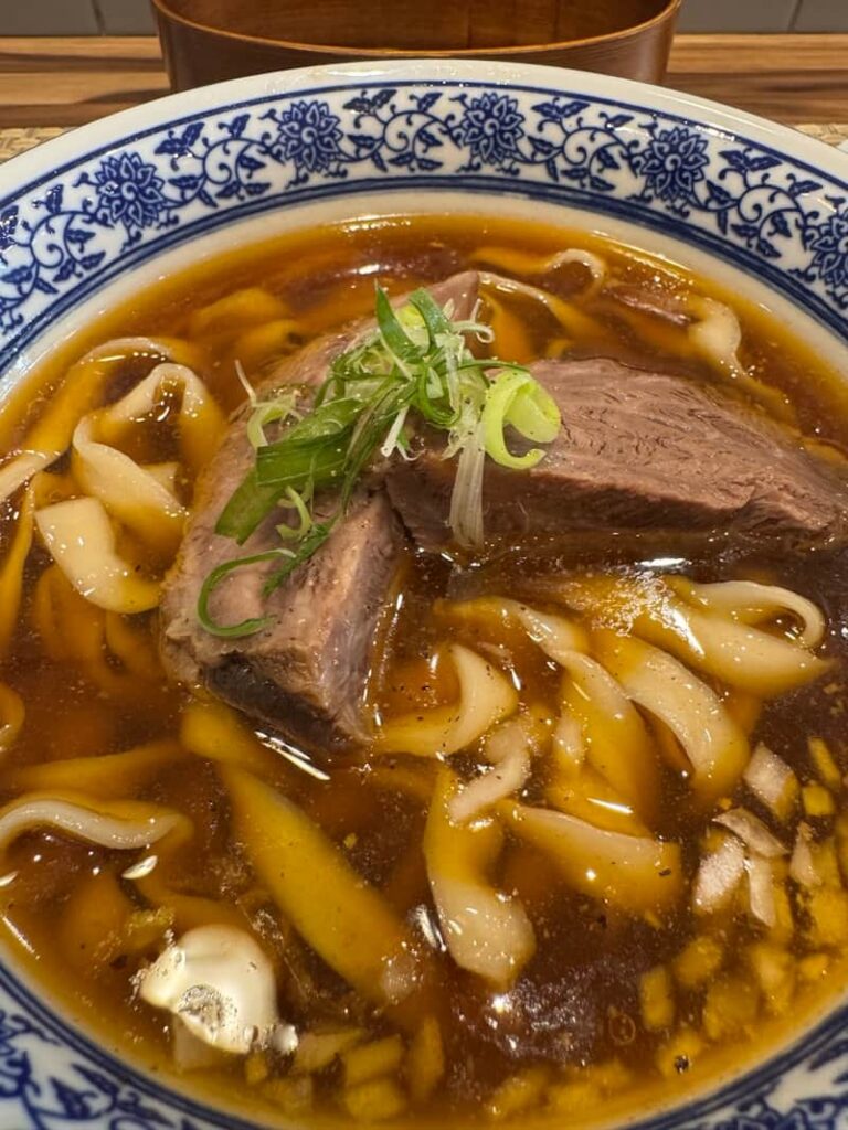 A rich, slow-simmered aromatic broth loaded with thick, chewy noodles, tender braised beef shank, and a garnish of fresh green onions.