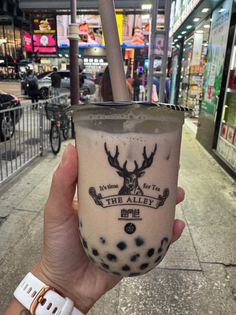 A hand holding a branded plastic cup of milk tea with dark tapioca pearls from The Alley, set against a bustling city street illuminated by evening lights.