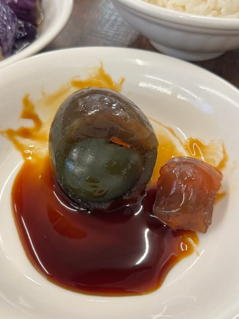 A peeled dark brown century egg (pidan) served in a white bowl with a savory soy-based dipping sauce.