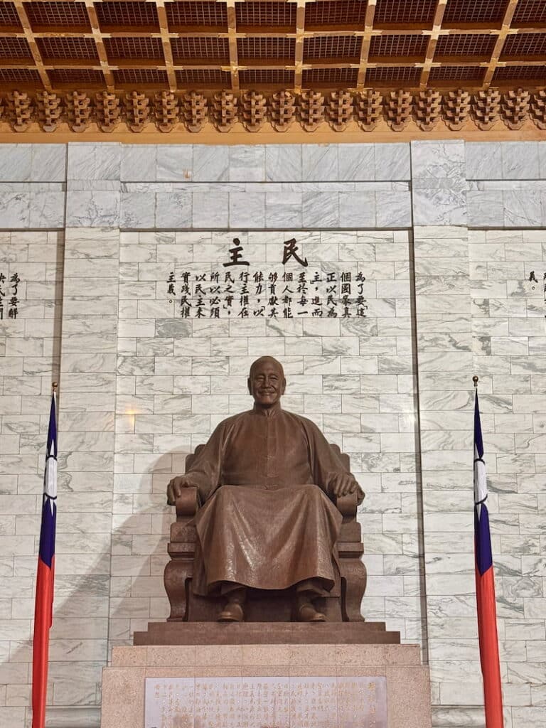 A bronze seated statue of Chiang Kai-shek inside the memorial hall, flanked by Taiwanese flags and set against a marble wall inscribed with Chinese text beneath an ornate golden ceiling.