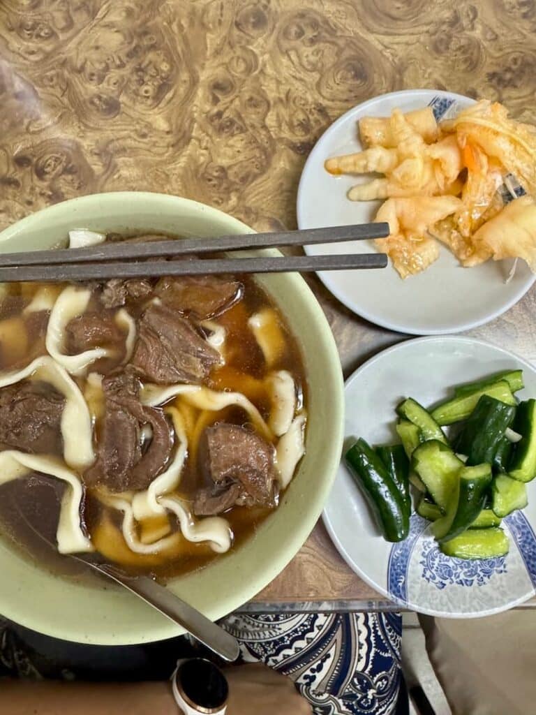 A comforting bowl of Taiwanese beef noodle soup with thick handmade noodles and tender beef shanks, served alongside small plates of pickled cucumbers and seasoned chicken feet.