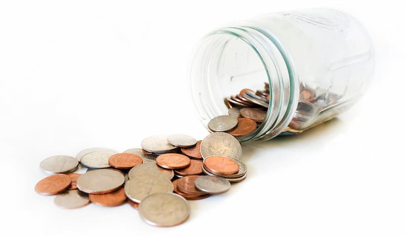 Coins spilling out of a glass jar tipped on its side against a light-colored background.