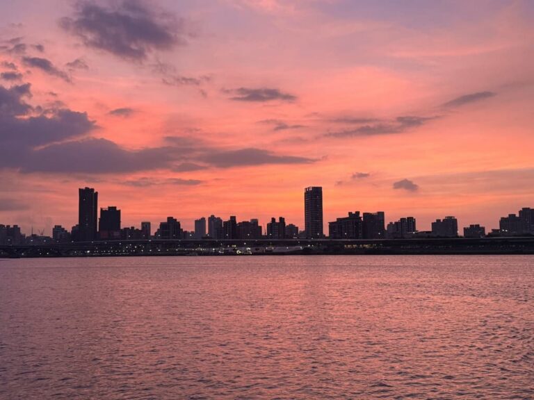 A vibrant pink and purple sunset casting a colorful reflection over the water with a distant Taipei city skyline silhouette along the horizon.