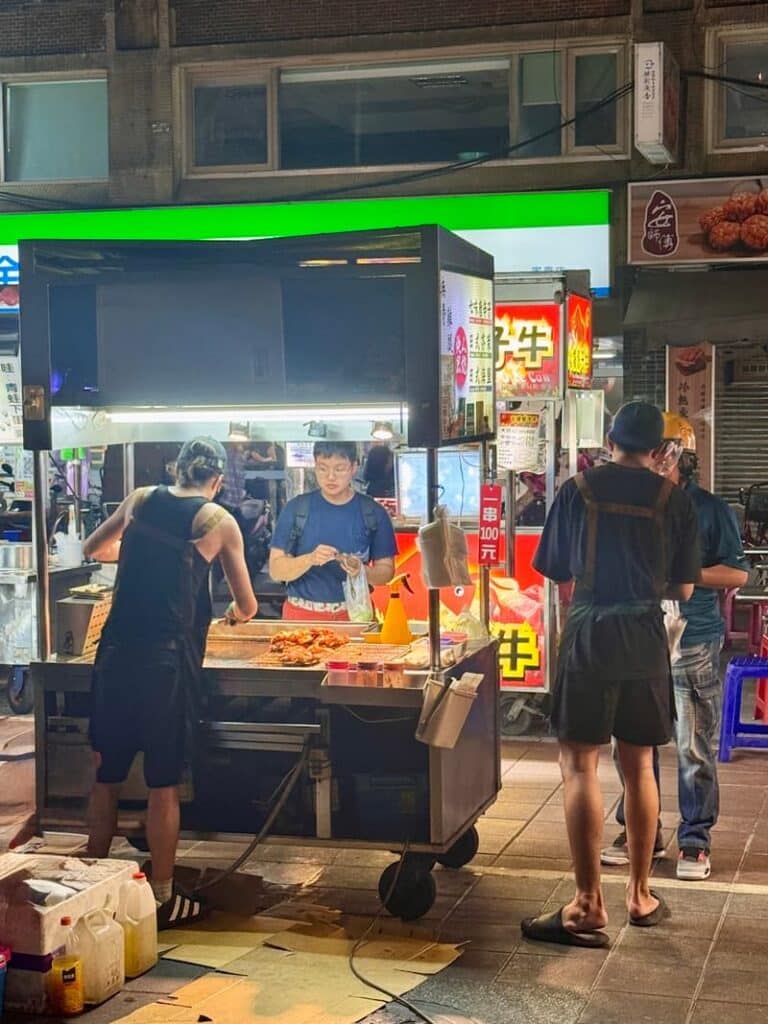 Vendors working at a brightly lit street food cart at night, with customers standing nearby.