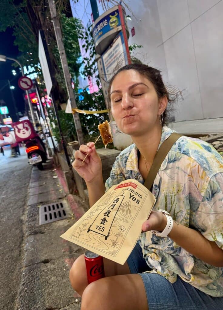 A traveler enjoying a bite of crispy fried chicken on a skewer while holding a paper takeout bag on a lit city street at night.