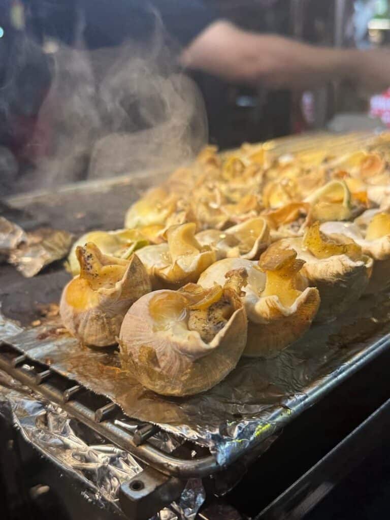 Fresh sea snails sizzling on a hot street food stall griddle, steaming with garlic and savory seasonings.