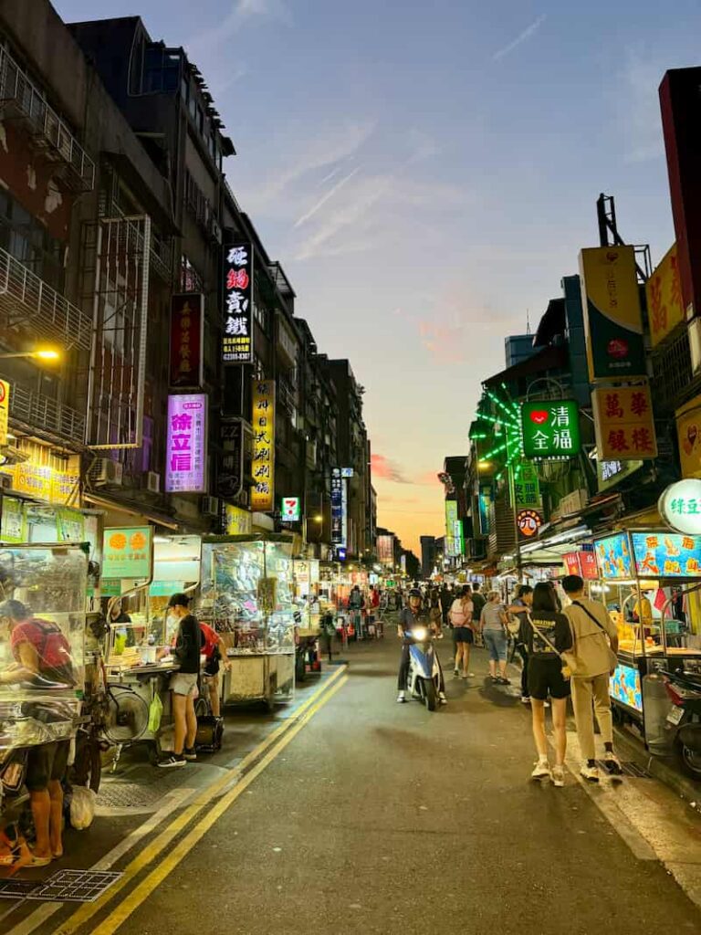 A narrow Taipei street market at dusk, lined with illuminated food stalls, glowing Chinese storefront signs, and pedestrians, with a scooter riding down the center lane.