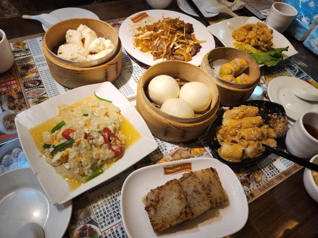 Table full of Cantonese dim sum, bamboo steamers, and stir-fried dishes at a Hong Kong eatery.