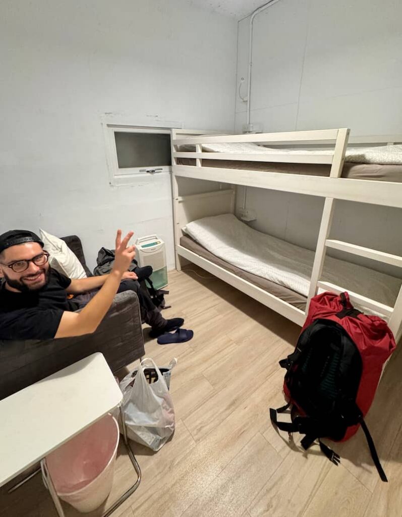 A smiling person wearing glasses and a cap, flashing a peace sign while seated in a hostel room in Ruifang, which features a white bunk bed, a red backpack on the floor, and a small window.