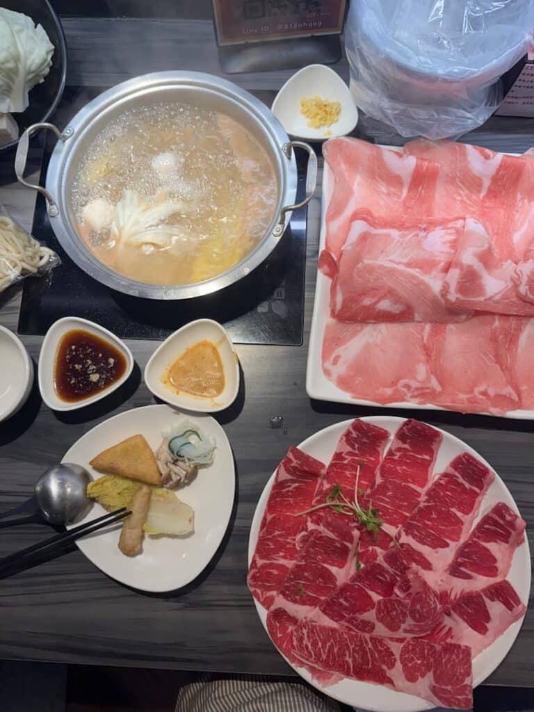 A tabletop hot pot spread featuring a boiling metal pot of broth, plates of thinly sliced raw beef and pork, side dishes, and dipping sauces.