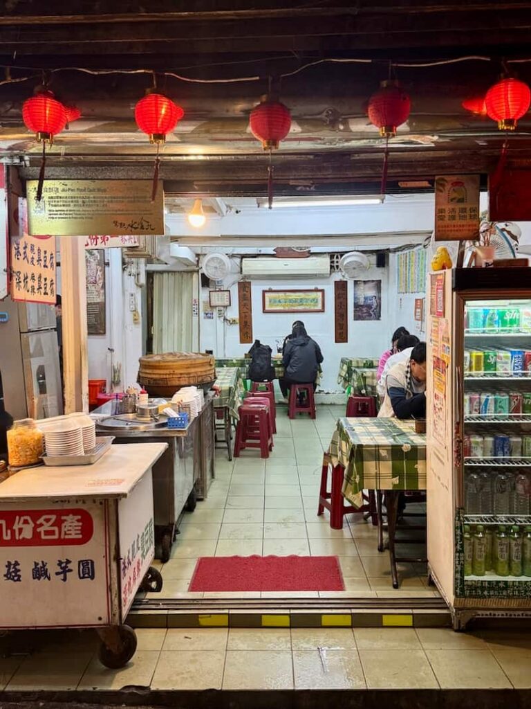 The interior and entrance of a small traditional restaurant adorned with hanging red lanterns, featuring checkered-cloth dining tables with patrons eating inside.