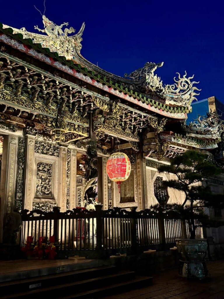 A traditional temple exterior illuminated at night, featuring ornate architectural carvings, detailed roof dragons, and a glowing red lantern, as featured in our Is Taipei worth visiting guide.