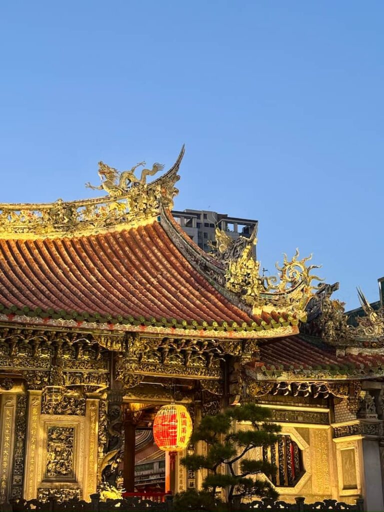 A detailed view of the temple's ornate tiled roof, golden dragon sculptures, and a glowing red lantern set against a clear blue sky.