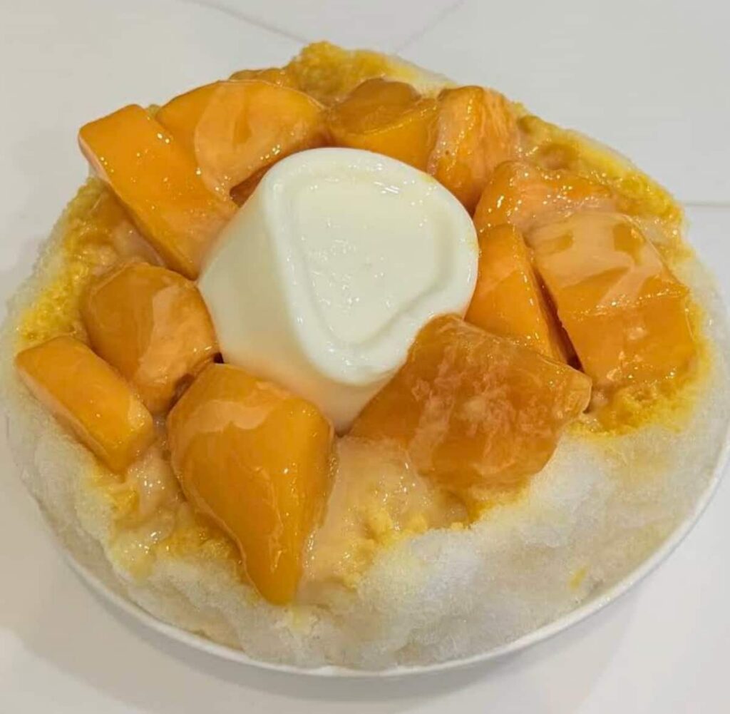 A bowl of fine shaved ice topped with chunks of bright orange mango and a smooth white milk pudding in the center.