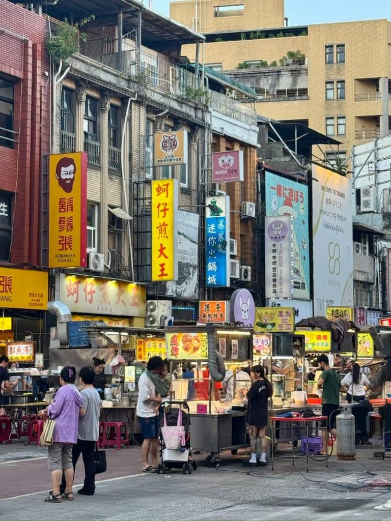 A busy Taipei street market scene with food stalls, pedestrians, and older multi-story buildings draped in colorful vertical Chinese signage under daylight.