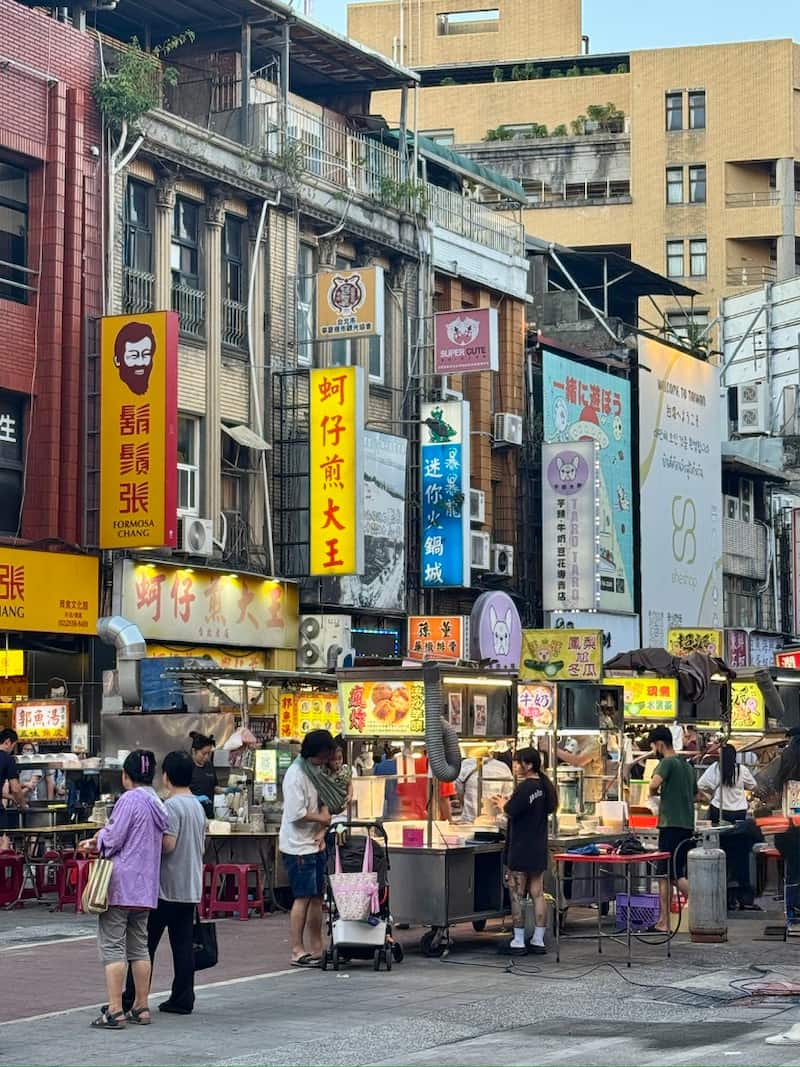A busy Taipei street market scene with food stalls, pedestrians, and older multi-story buildings draped in colorful vertical Chinese signage under daylight.