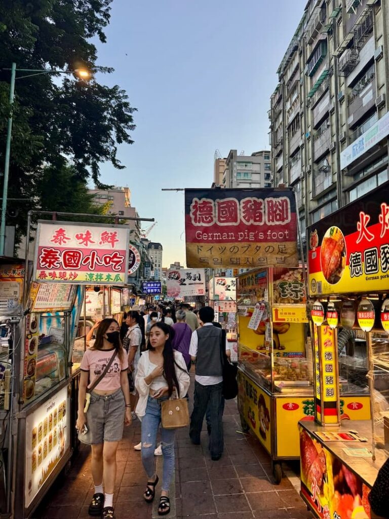 Two young women walking through a crowded Taipei street market lined with food vendors, featuring prominent signs in multiple languages including a banner for German pork knuckles.