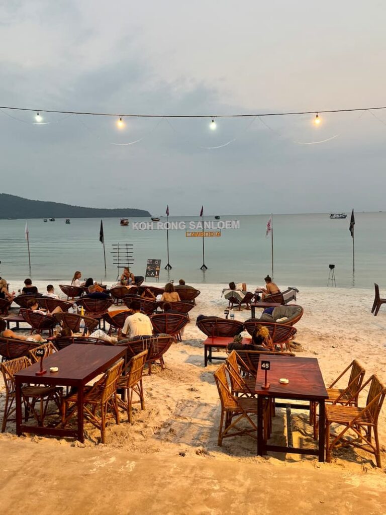 Open-air beach restaurant setup with wooden tables and chairs on the sand at Saracen Bay, Koh Rong Sanloem, with string lights overhead and guests relaxing overlooking the ocean.