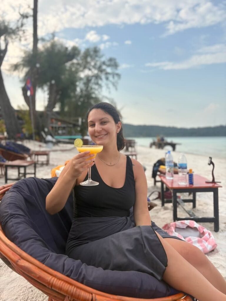 The travel blogger holding a tropical cocktail garnished with a lime on a lounge chair on the white sand at Saracen Bay, Koh Rong Sanloem.