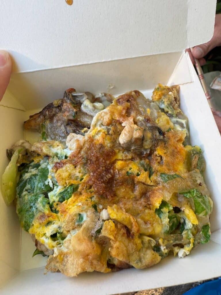A takeout container filled with a traditional Taiwanese oyster omelette featuring eggs, plump oysters, and leafy greens, as featured in our night markets in Taipei guide.