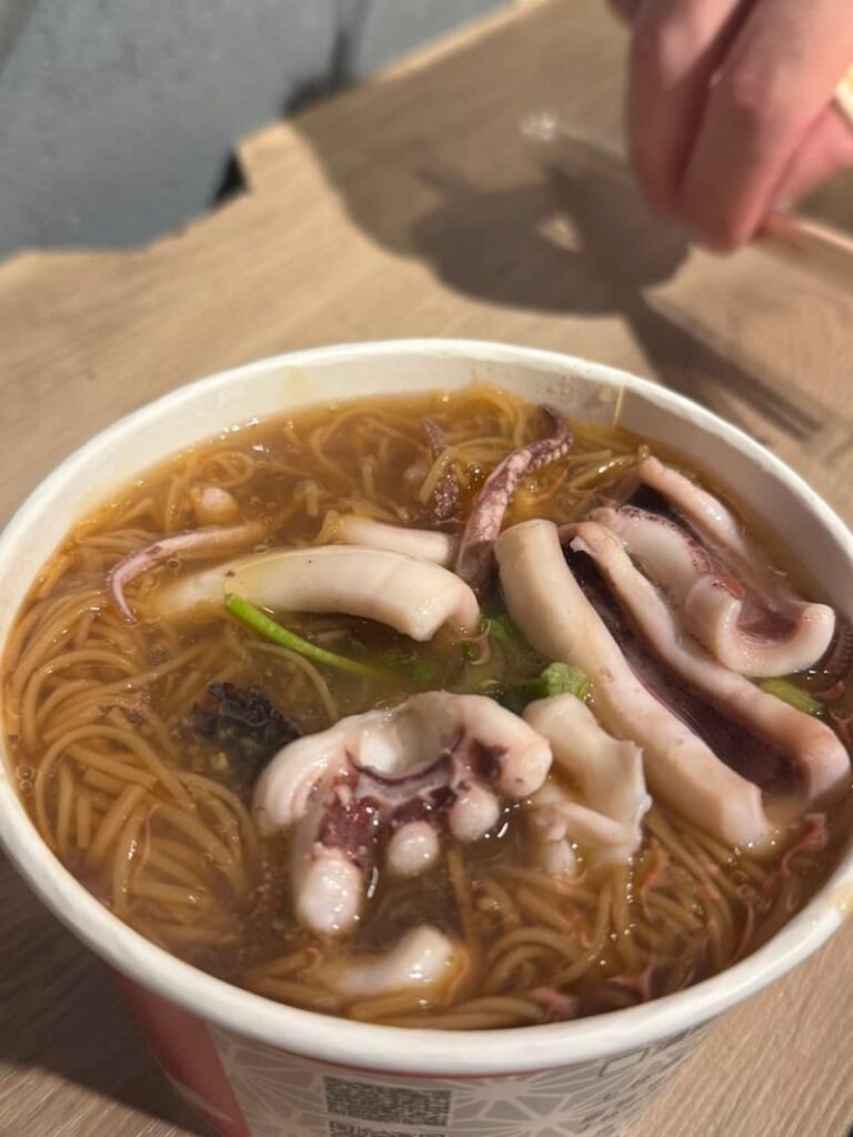 A takeaway paper bowl filled with thick, brown vermicelli noodles in a rich, umami broth topped with tender squid and octopus tentacles.