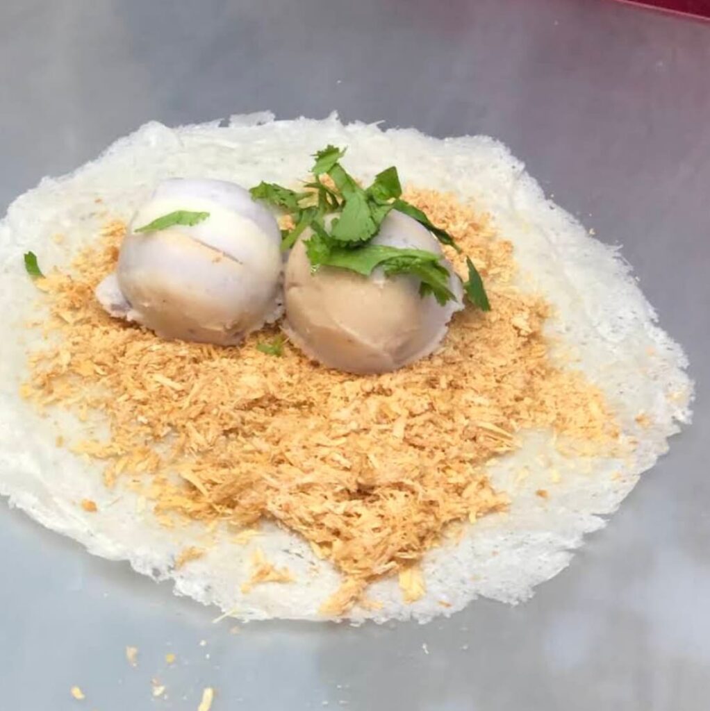 A thin white spring roll wrapper laid flat, generously covered with shaved peanut brittle, two scoops of ice cream, and garnished with fresh cilantro leaves.