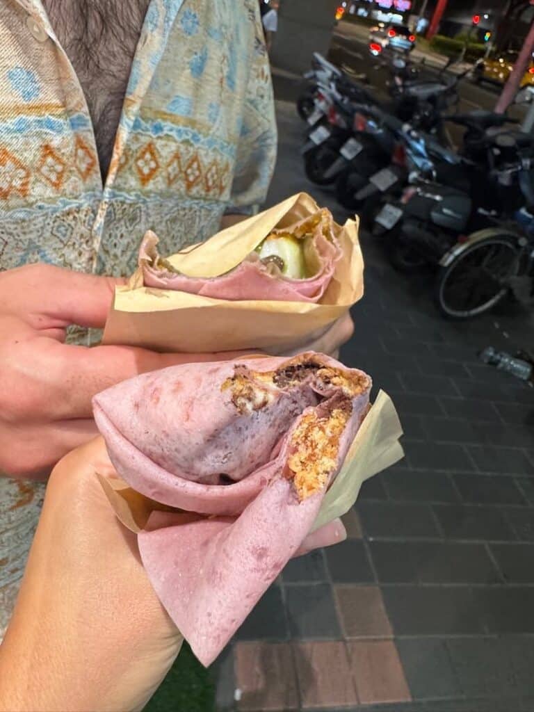 Two people holding Taiwanese peanut ice cream rolls wrapped in pink spring roll wrappers and paper sleeves on a street.