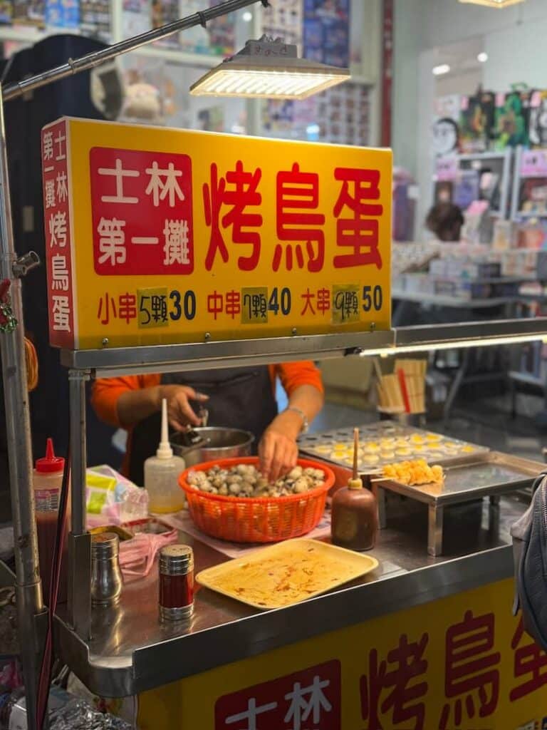 A bright yellow and red street food stall sign reading "烤鸟蛋" (grilled quail eggs) above a vendor preparing skewers of quail eggs on a flat griddle.