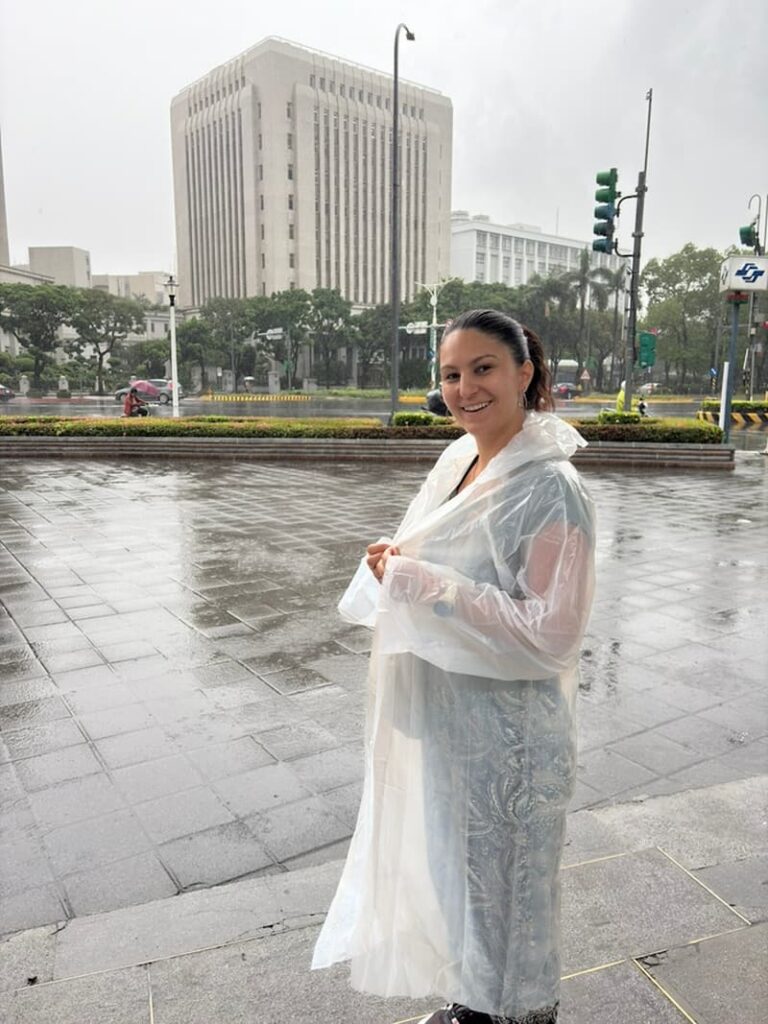 A smiling person wearing a clear plastic disposable raincoat on a wet city plaza during a rainy day in Taipei.