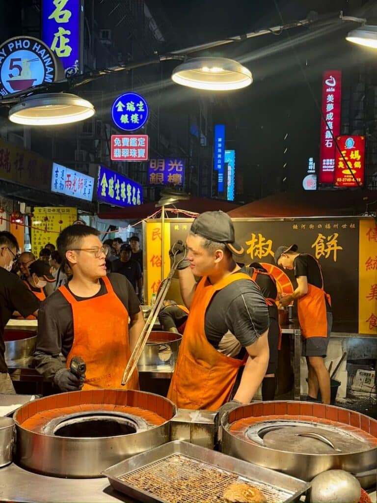 Two vendors wearing orange aprons working at a busy Raohe Night Market stall with traditional cylindrical baking ovens.