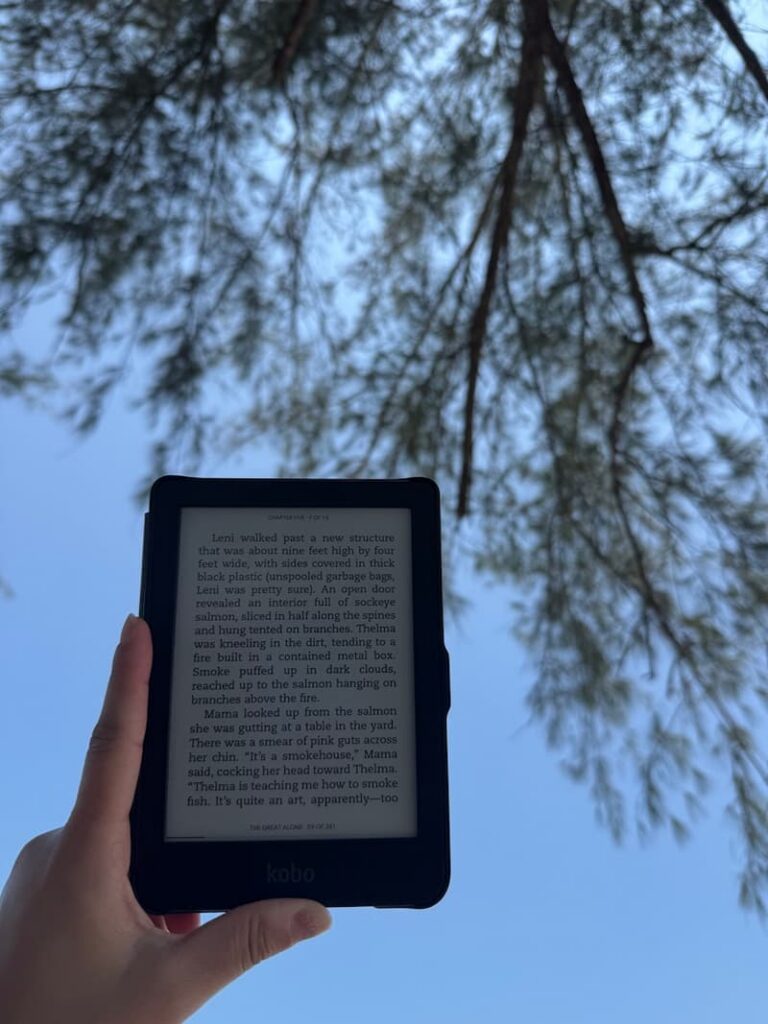 Person holding a Kobo e-reader outdoors against a backdrop of trees and a blue sky.