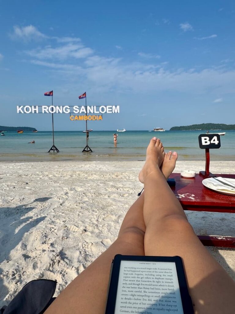 the travel blogger relaxing on the white sand at Saracen Bay, Koh Rong Sanloem, reading an e-reader with bare feet propped up and turquoise water in the background, as featured in our how to spend 2 days in koh rong samloem guide.