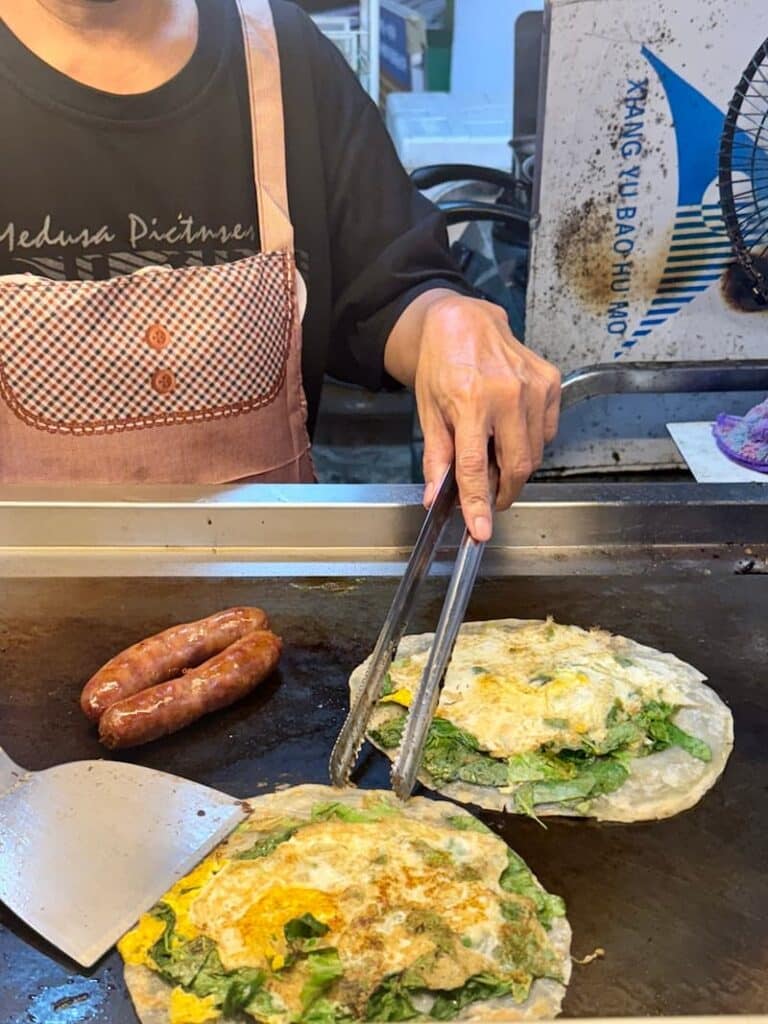 A vendor using metal tongs to prepare Taiwanese egg crepes with fresh greens alongside sizzling sausages on a flat street food griddle.