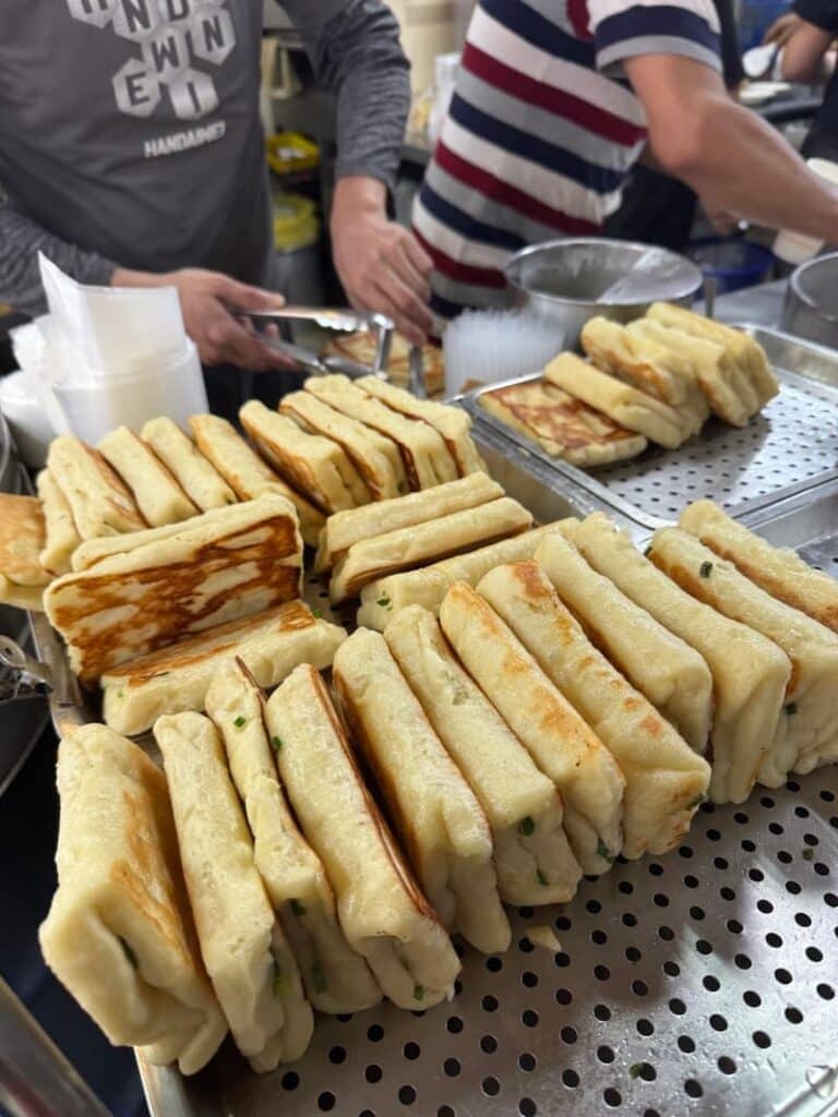 Stacks of freshly cooked, golden-brown folded Taiwanese egg crepes (danbing) arranged on metal trays at a busy breakfast stall, with vendors working in the background.