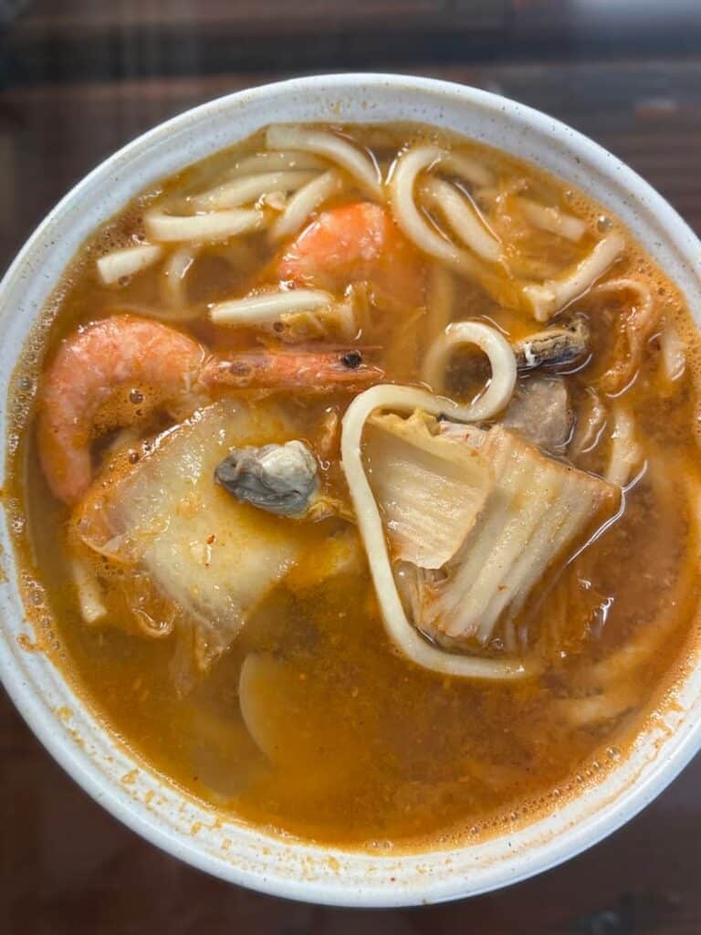 A white bowl filled with a savory seafood noodle soup containing plump shrimp, oysters, cabbage, and thick white noodles.