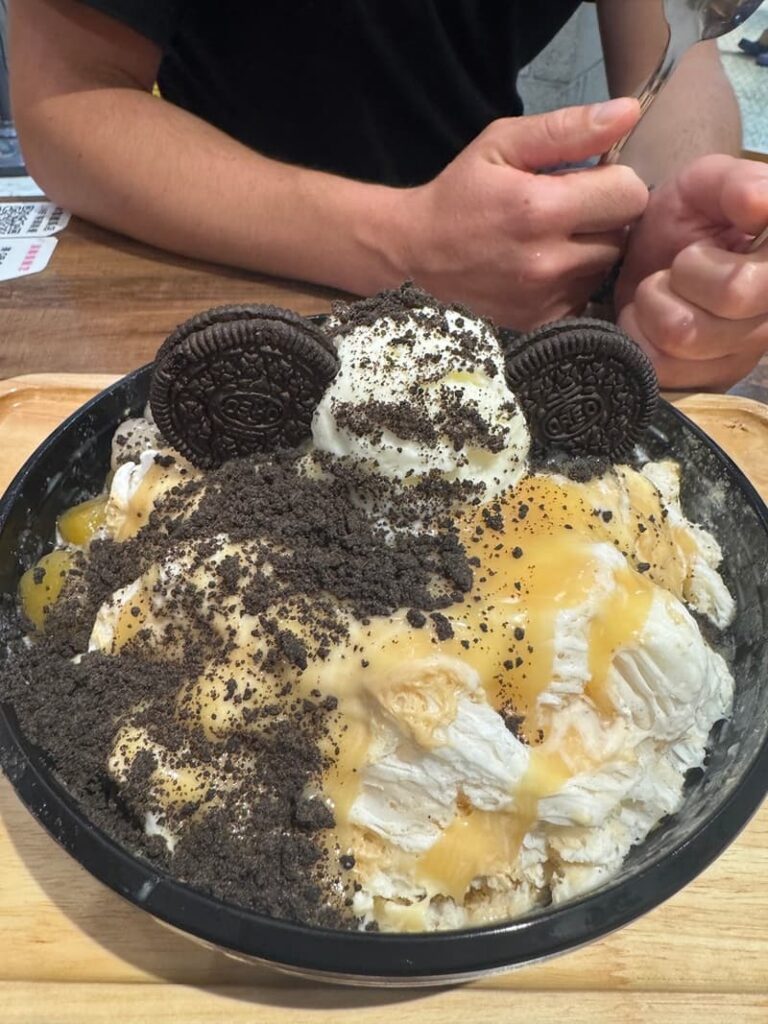 A decadent bowl of shaved ice topped with vanilla ice cream, whole Oreo cookies, crushed cookie crumbs, and caramel drizzle.