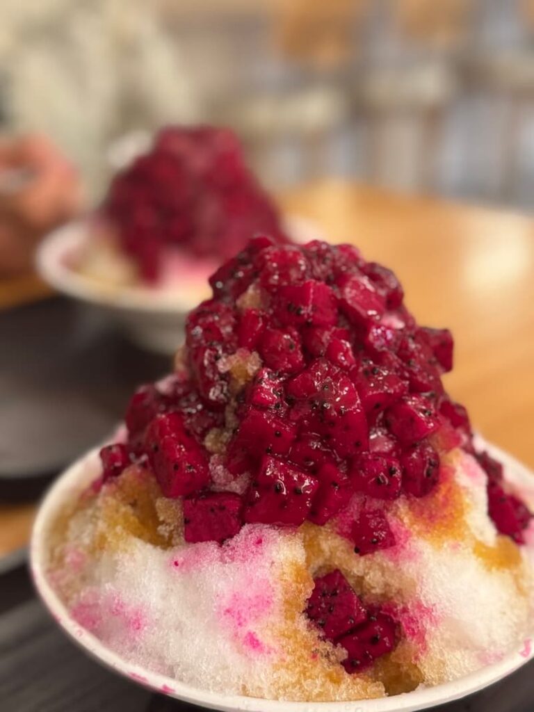 A towering bowl of Taiwanese shaved ice covered in bright pink chunks of fresh dragon fruit and sweet syrup.