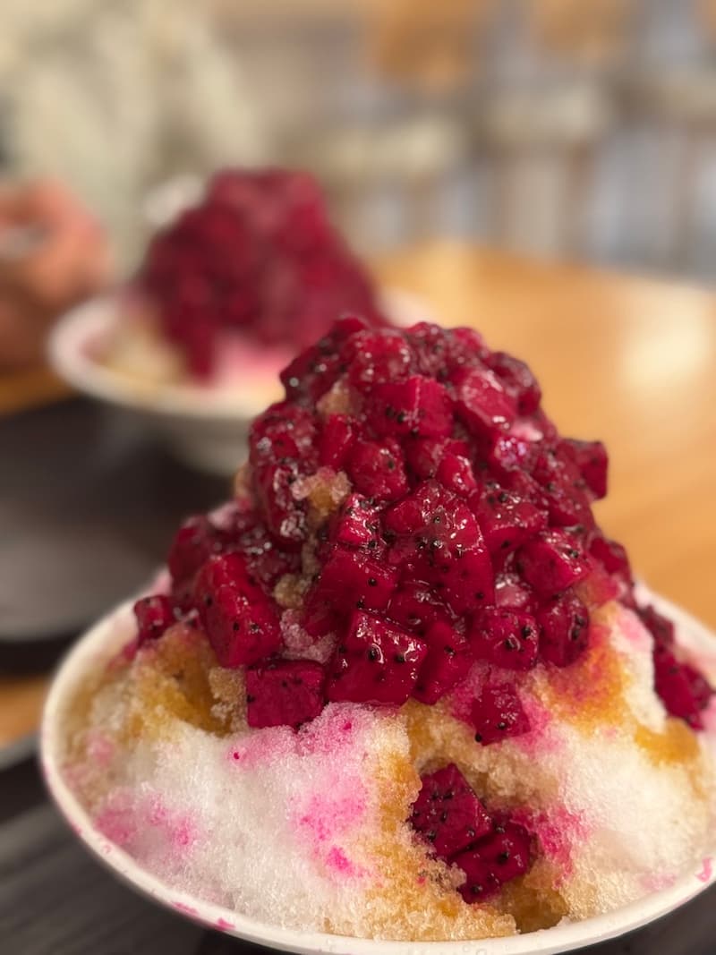 A towering bowl of Taiwanese shaved ice covered in bright pink chunks of fresh dragon fruit and sweet syrup.