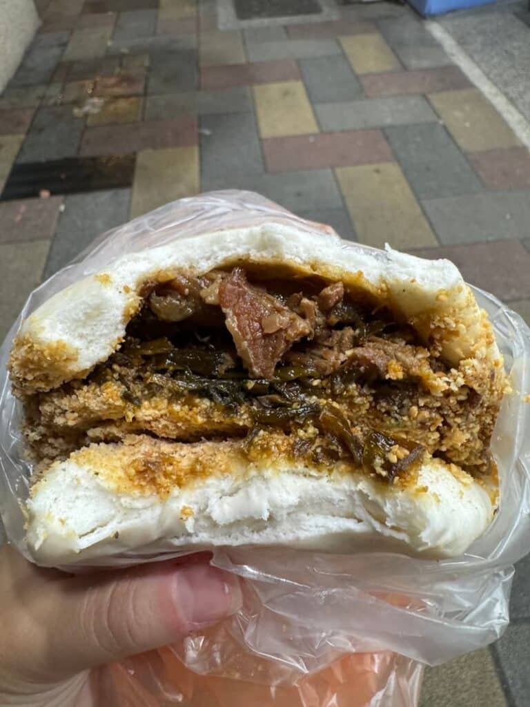 A close-up of a large, fluffy white steamed bun torn open to reveal a generous filling of savory minced meat and dark preserved greens.