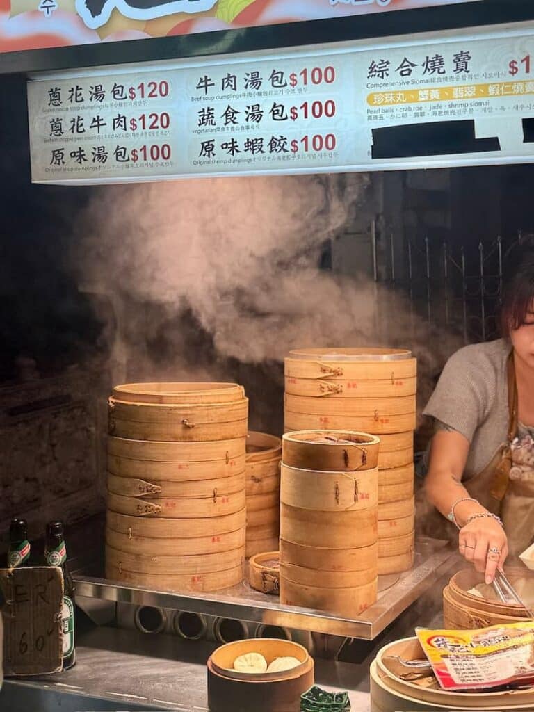 A bustling street food stall displaying towering stacks of steaming bamboo steamers underneath an illuminated menu board, as seen in our what to eat in Taipei guide.