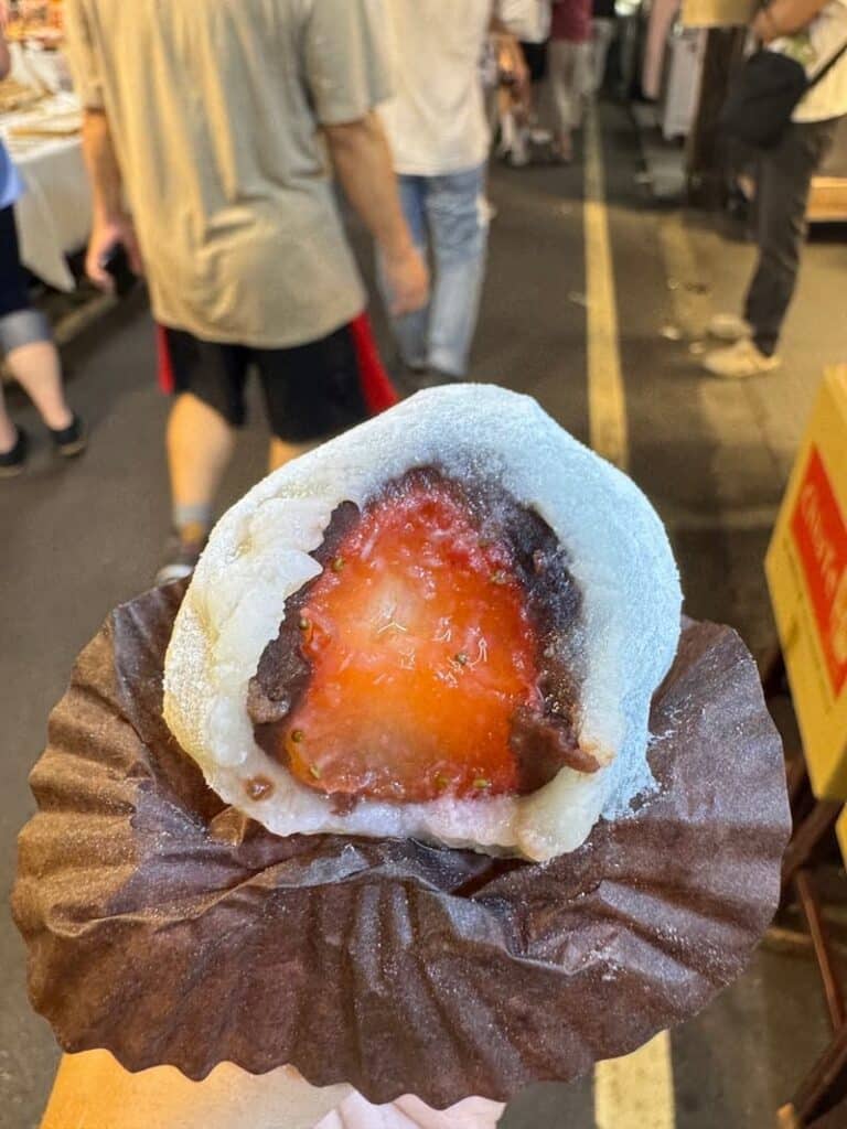 Close-up of a strawberry daifuku mochi cut in half, revealing a fresh red strawberry wrapped in sweet red bean paste and soft white mochi skin at a night market.
