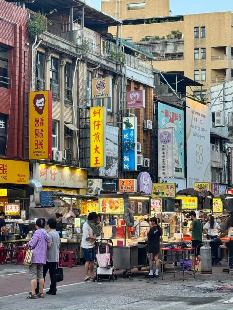 A bustling Taipei street scene featuring colorful vertical storefront signs, historic multi-story buildings, and active food vendor stalls at ground level.