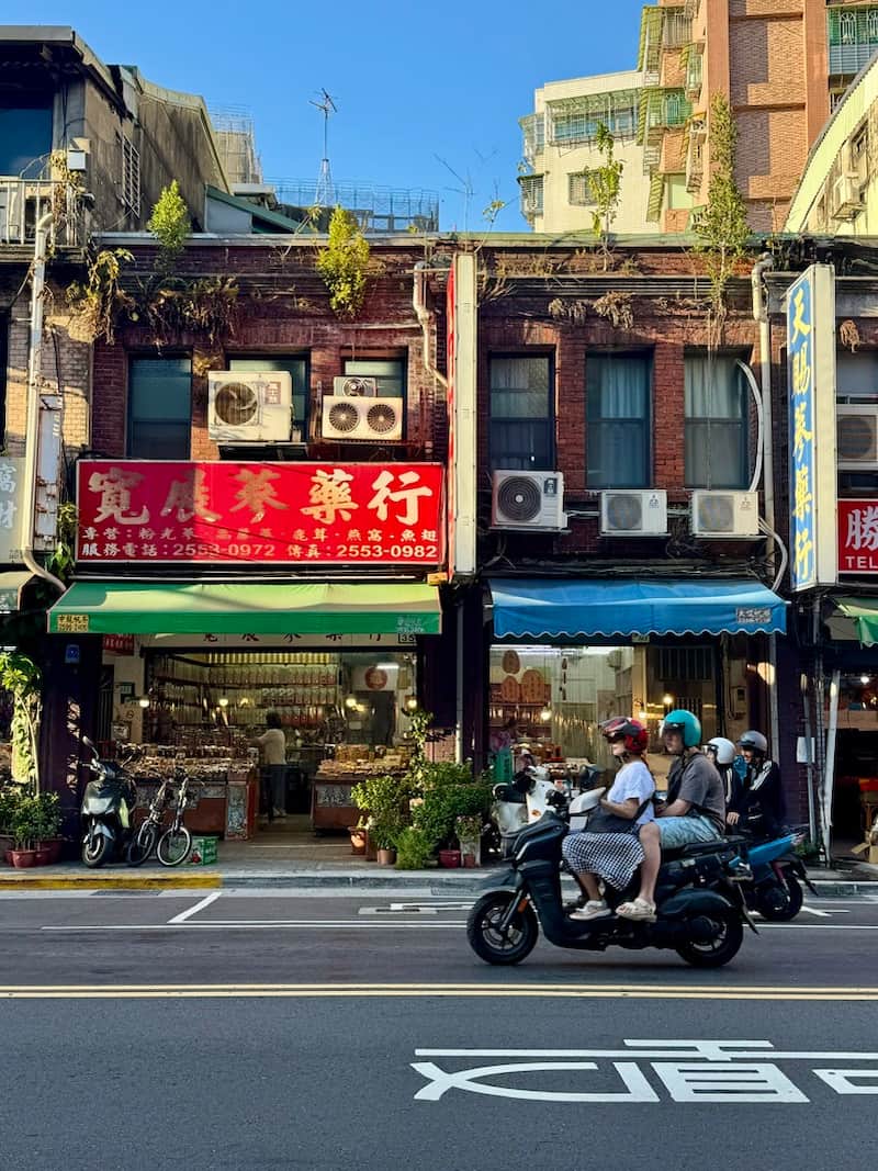 People riding a scooter past traditional shops with red Chinese signs on a Taipei street, as featured in our guide about the best esims in Taiwan.