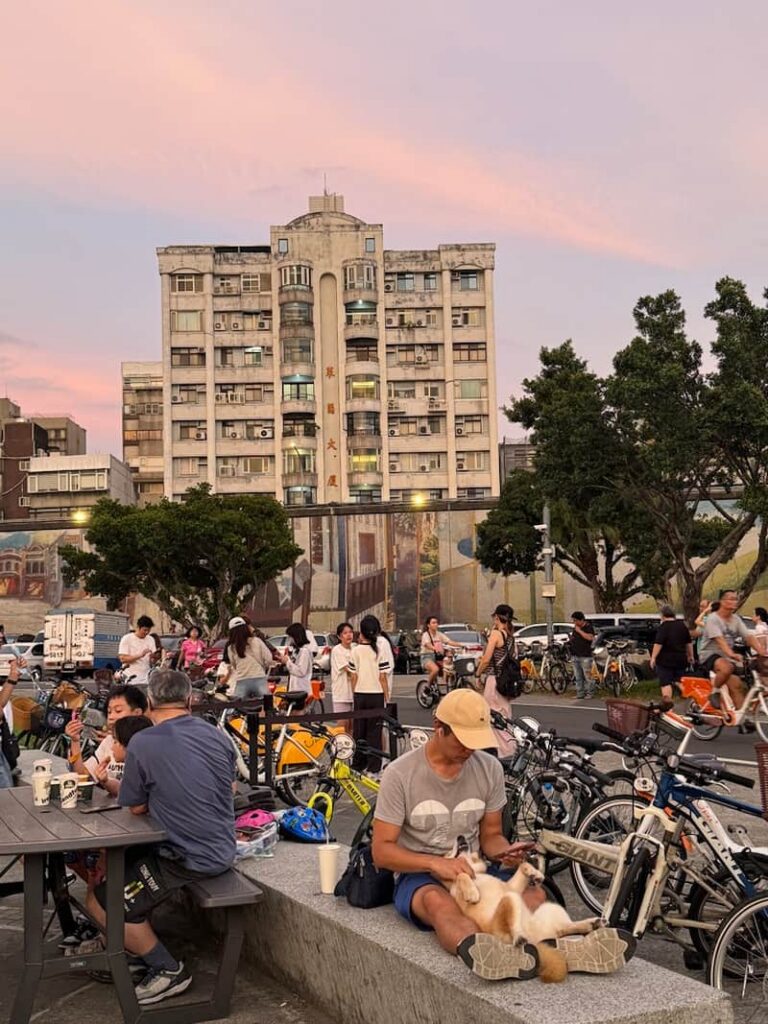 People relaxing outdoors in Taipei at dusk, featuring parked bicycles and a dog resting on someone's lap.