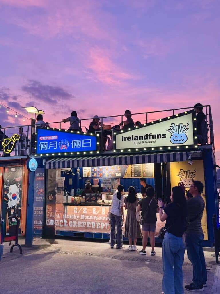 A colorful shipping container food vendor named "irelandfuns" at dusk under a vibrant purple and pink sunset sky, with patrons ordering at the counter and relaxing on the rooftop seating area.