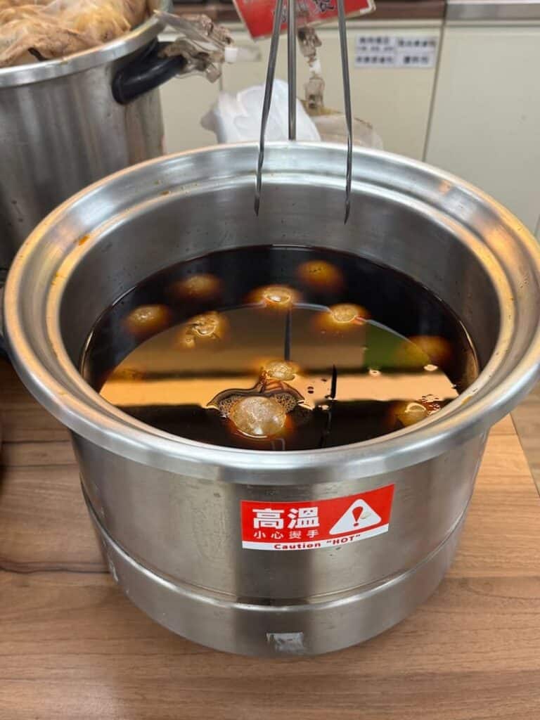 A large stainless steel pot filled with dark, bubbling herbal broth and simmering tea eggs, with a "Caution HOT" warning label.