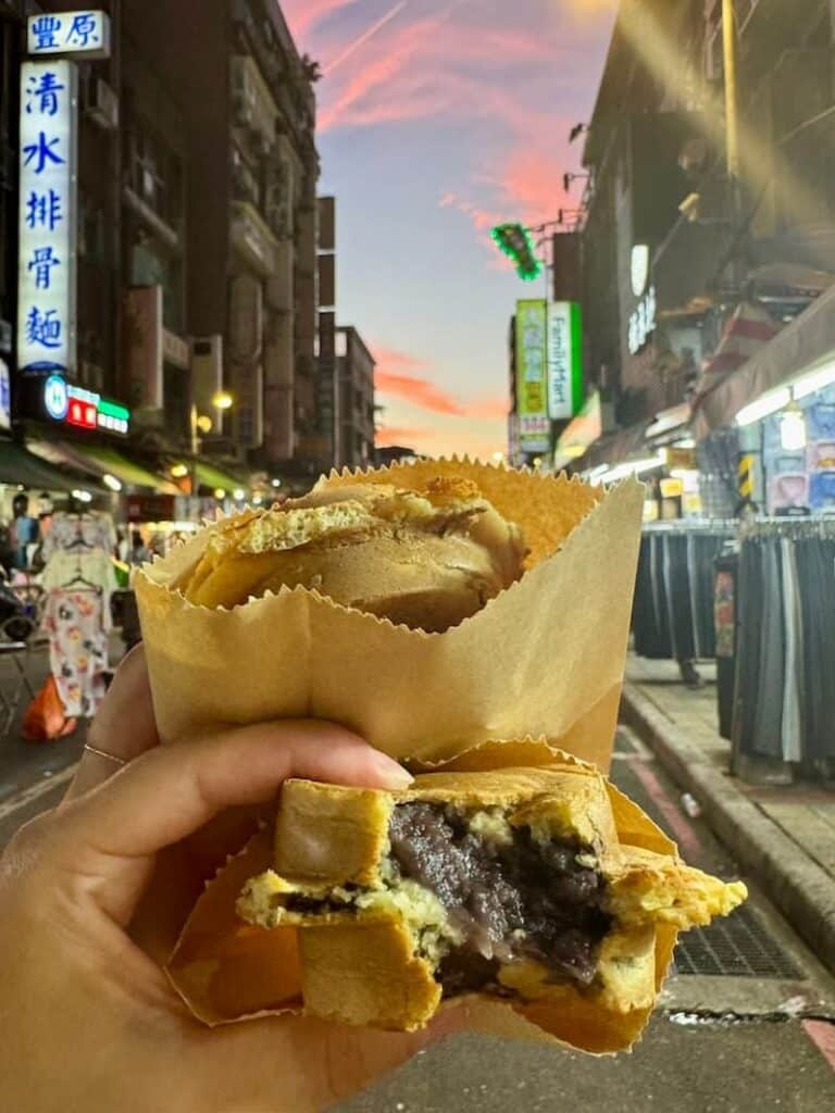 A crispy Taiwanese wheel cake (Taipei night market pastry) broken open to reveal a thick red bean paste filling, held against a vibrant sunset street background.